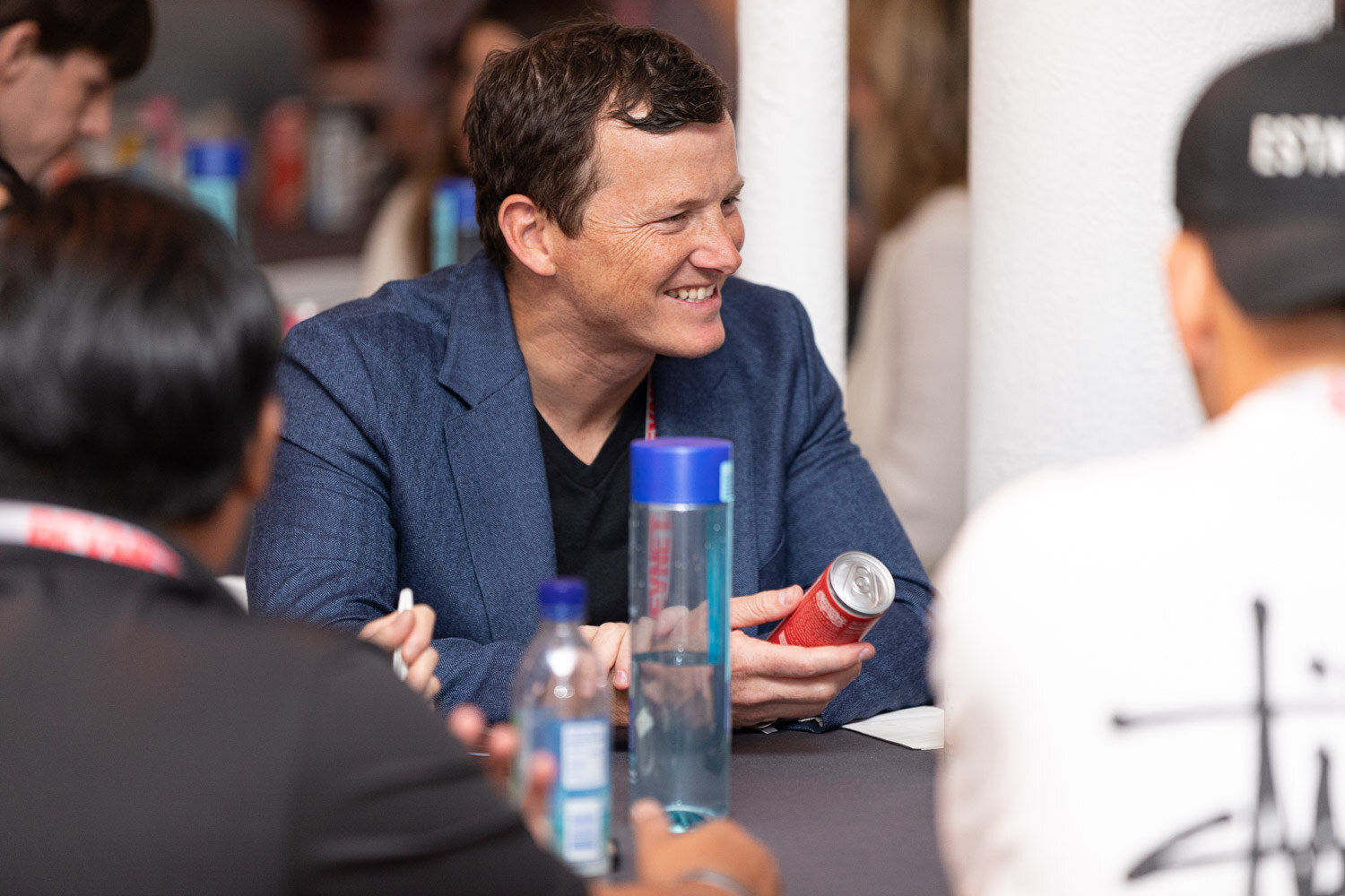 A smiling man in a navy blazer sits at a table with a water bottle and a red can in front of him while others talk around him.