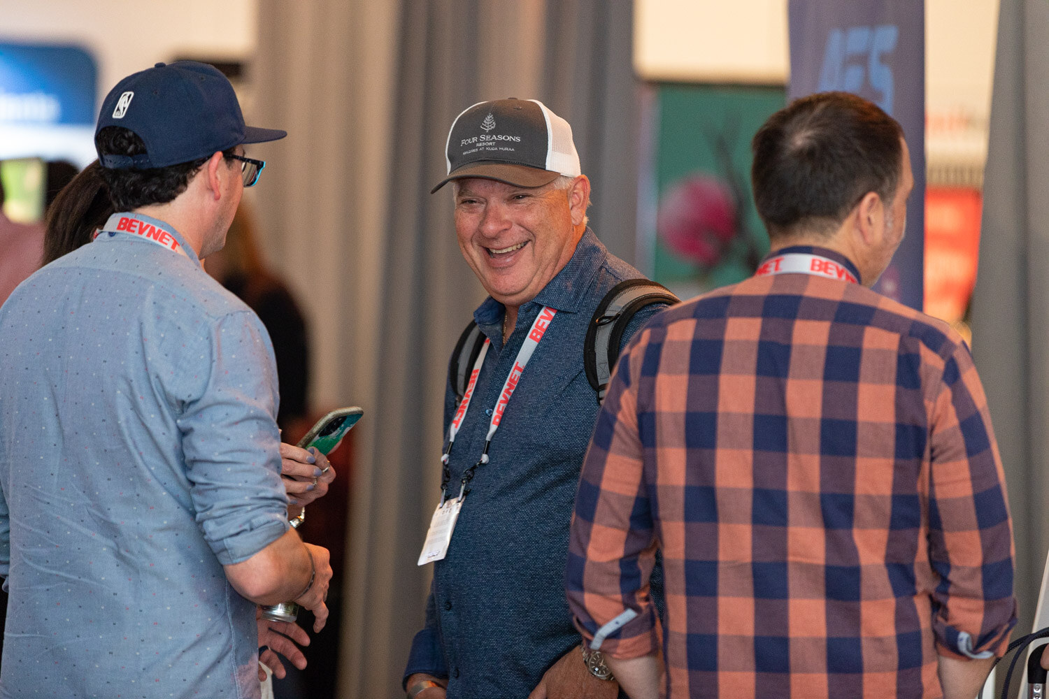 Three men with lanyards stand together in a loft space, one laughing in a mesh-back cap, with sponsor banners behind them.