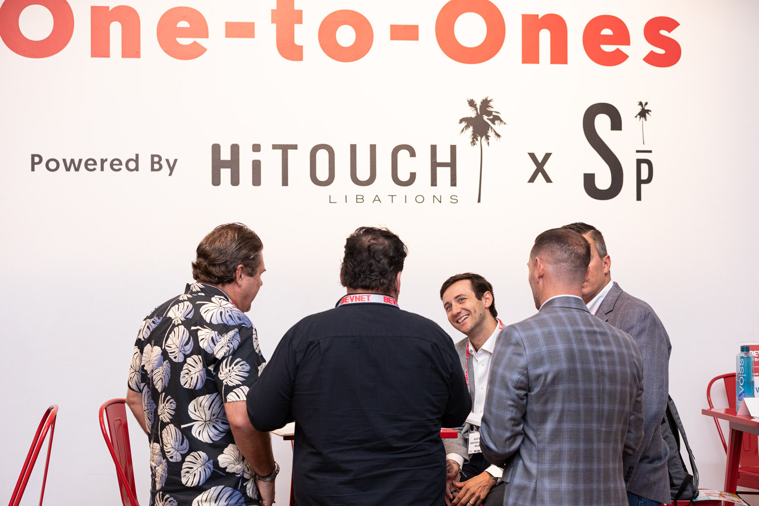 Four men in jackets and a patterned shirt stand together smiling, with a One-to-Ones sign powered by Hi-Touch Libations on the wall behind them.