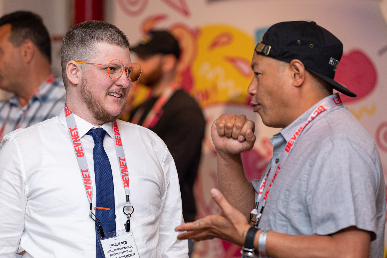 A man in a white shirt and navy tie and a man in a black cap and gray shirt gesture while talking, both with BevNET lanyards, in front of a colorful booth.