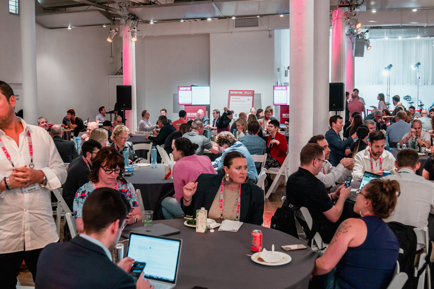 Wide view of many attendees with lanyards seated at round gray tables talking, eating and using laptops in a loft room.