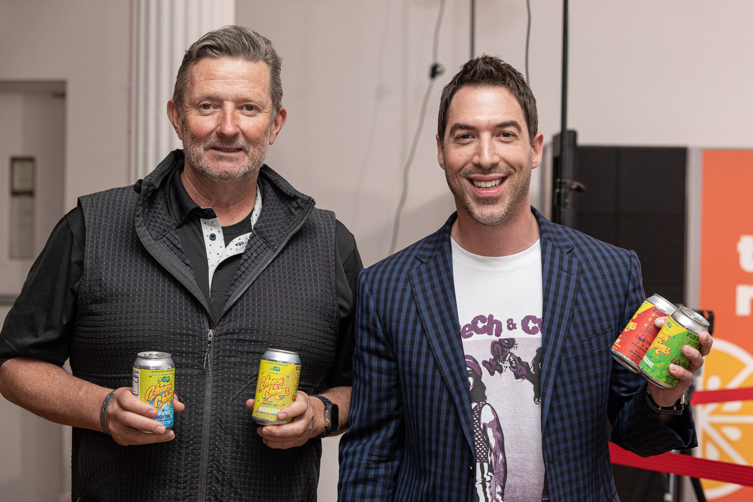 A man in a black vest and a man in a patterned navy blazer smile at the camera, each holding a yellow can.