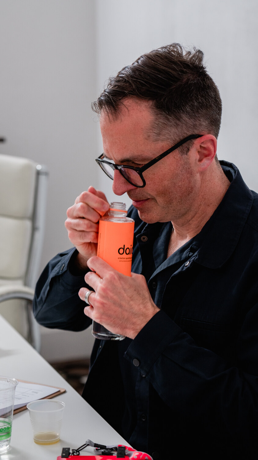 A man with glasses in a dark shirt holds an orange-labeled bottle up to his nose in a bright room.