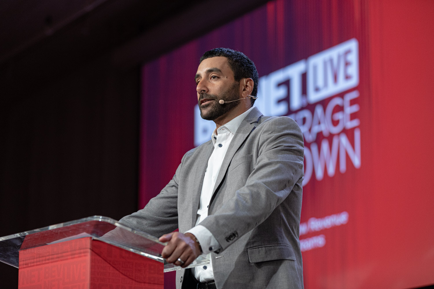 A man in a gray blazer and white shirt with a headset microphone speaks at a clear podium in front of a red BevNET Live screen.