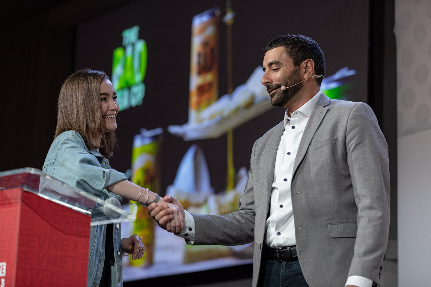 A woman in a denim jacket shakes hands with a man in a gray blazer at a clear podium with an illustrated slide behind them.