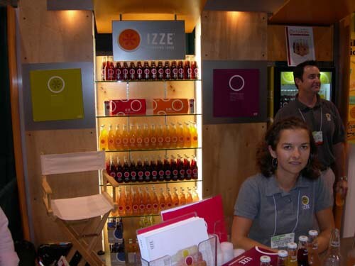A woman in a gray polo sits at a table while a man stands behind her, in front of lit shelves of IZZE bottles under an IZZE sign with colored square panels on either side.