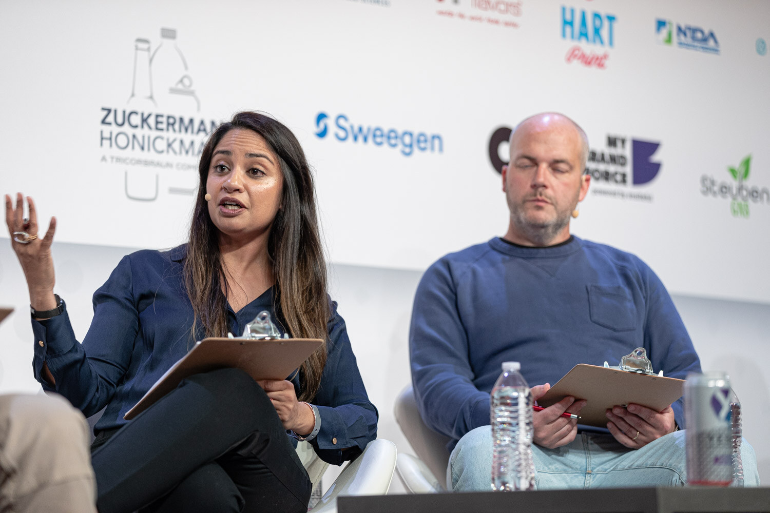 A woman in a navy top gestures while seated next to a bald man in a blue sweater, both holding clipboards in front of a sponsor wall.