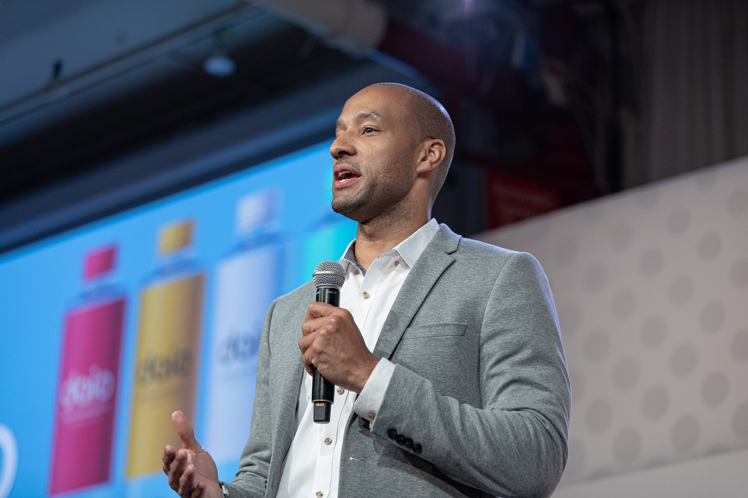 A bald man in a gray blazer and white shirt speaks into a microphone in front of a blue slide showing colorful bottles.