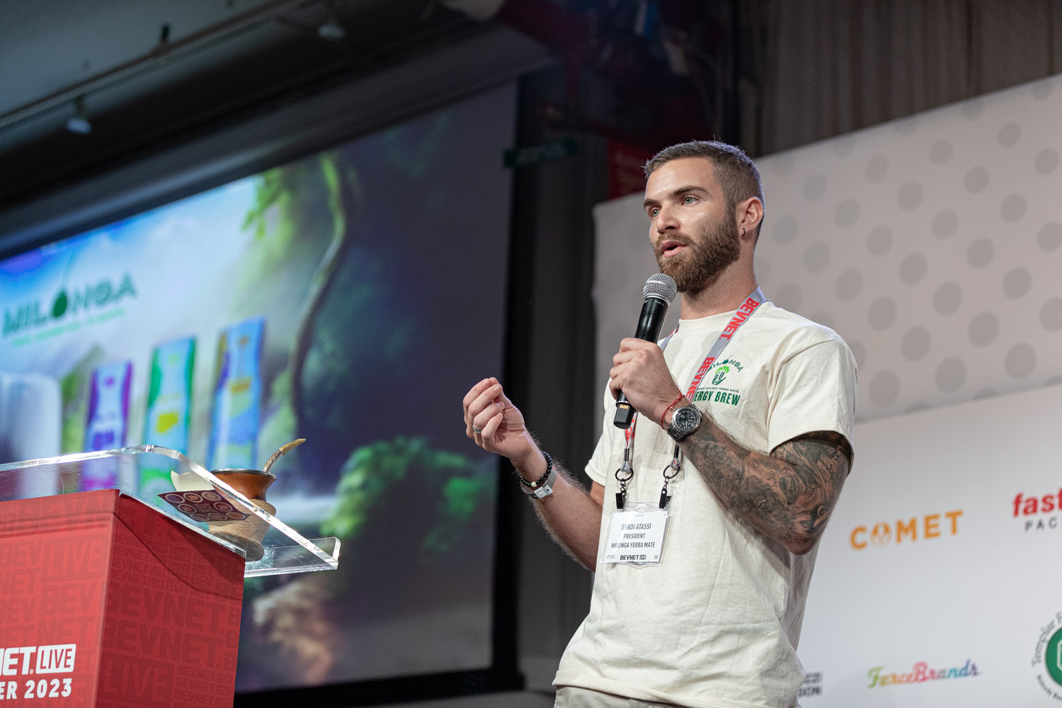A tattooed man in a cream T-shirt and lanyard speaks into a microphone beside a red podium, with a slide reading Milonga behind him.