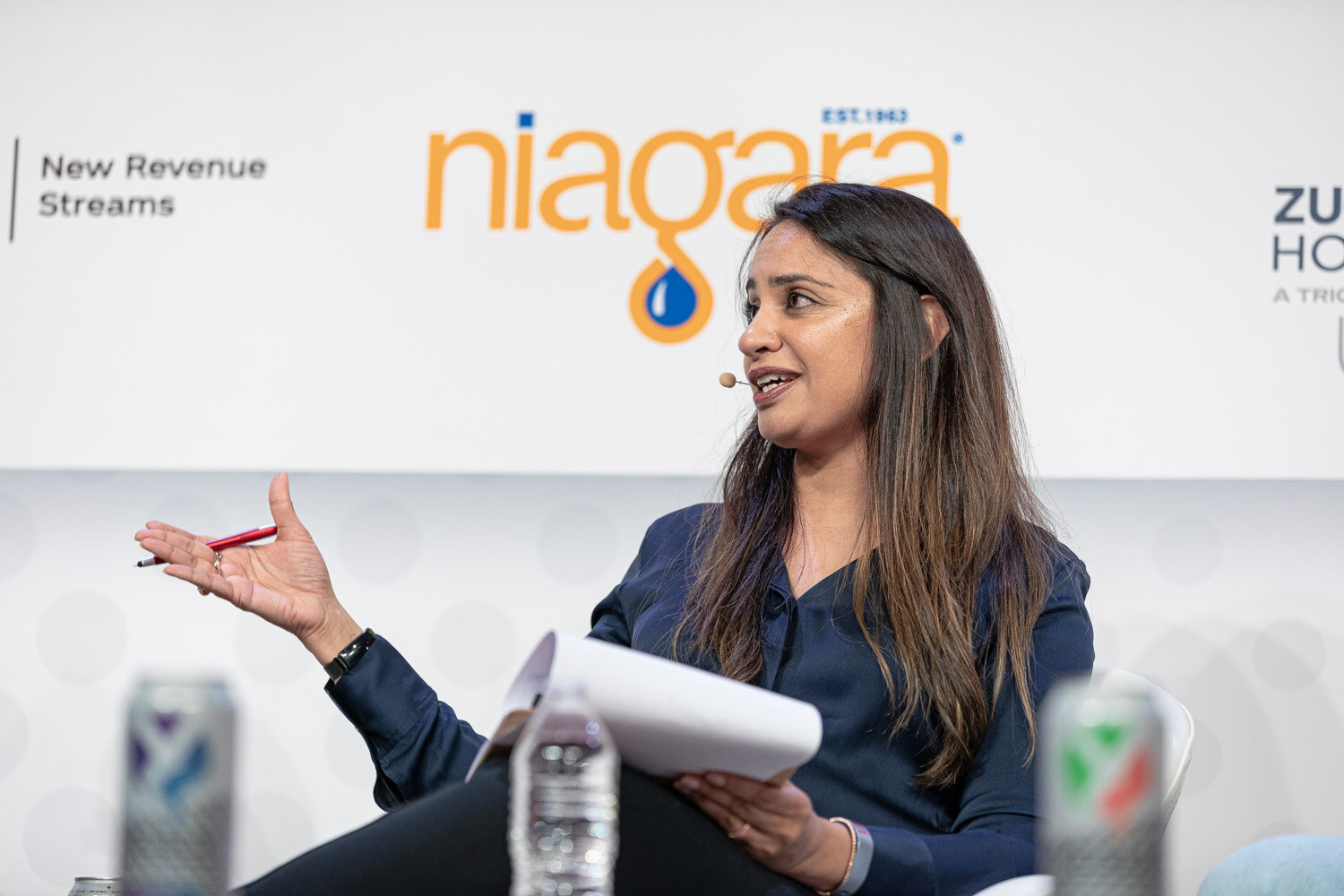 A woman with long dark hair in a navy top holds a pen and rolled notes while speaking, with a Niagara sign behind her.