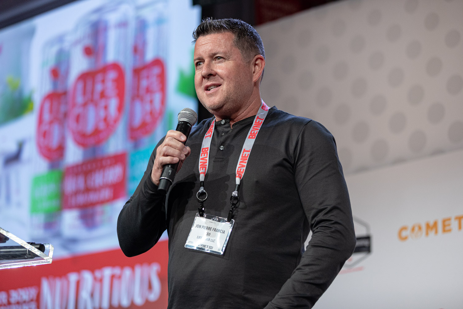 A man in a black shirt with a lanyard speaks into a microphone in front of a slide showing cans with the word Nutritious.