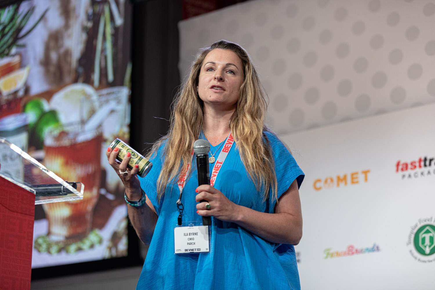 A woman with long blonde hair in a bright blue top holds a small can and a microphone with a slide of a cocktail behind her.