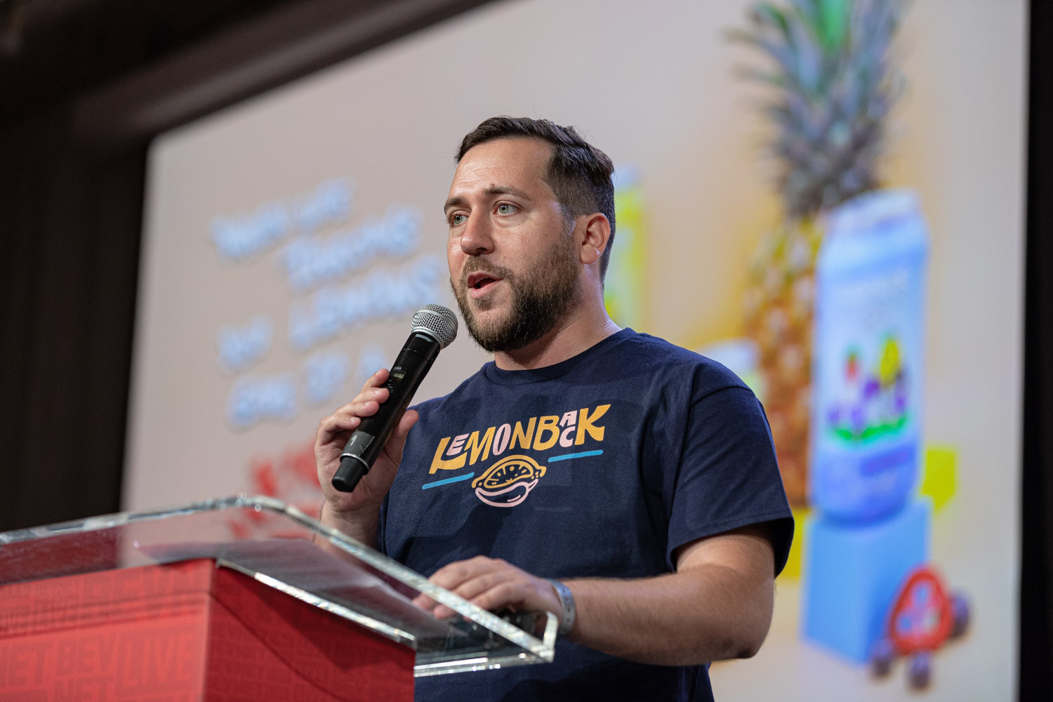 A bearded man in a navy T-shirt with a yellow logo speaks into a microphone in front of a slide with pineapple and a blue can.