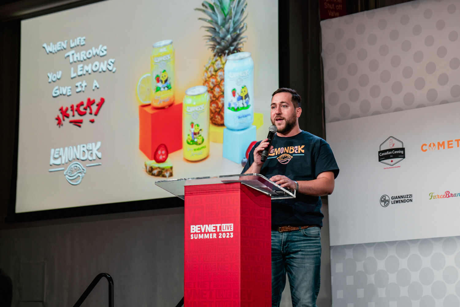 A man in a dark T-shirt and jeans stands at a red BevNET Live podium with a slide of pineapple and lemon drinks and a lemon-themed slogan behind him.