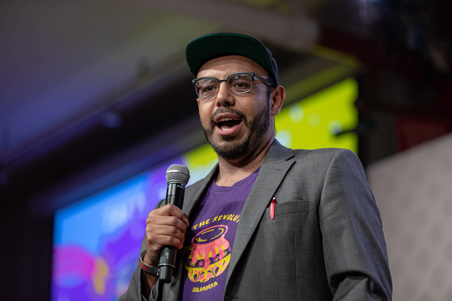 A man with glasses, a dark cap and a purple graphic T-shirt under a gray blazer speaks into a microphone in front of a bright slide.