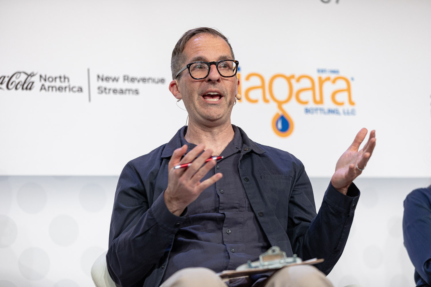 A man with glasses in a dark button-up shirt holds a pen and gestures with both hands in front of a Coca-Cola North America New Revenue Streams and Niagara sign.