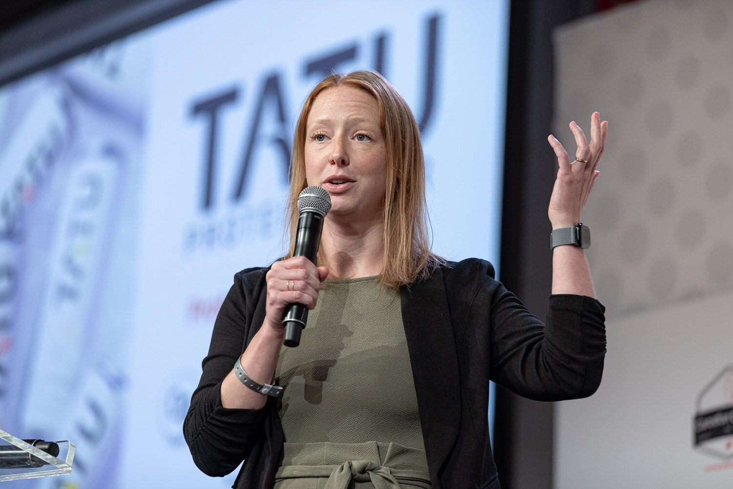 A woman in a black cardigan and olive top holds a microphone and raises one hand in front of a large slide that reads Tatu.