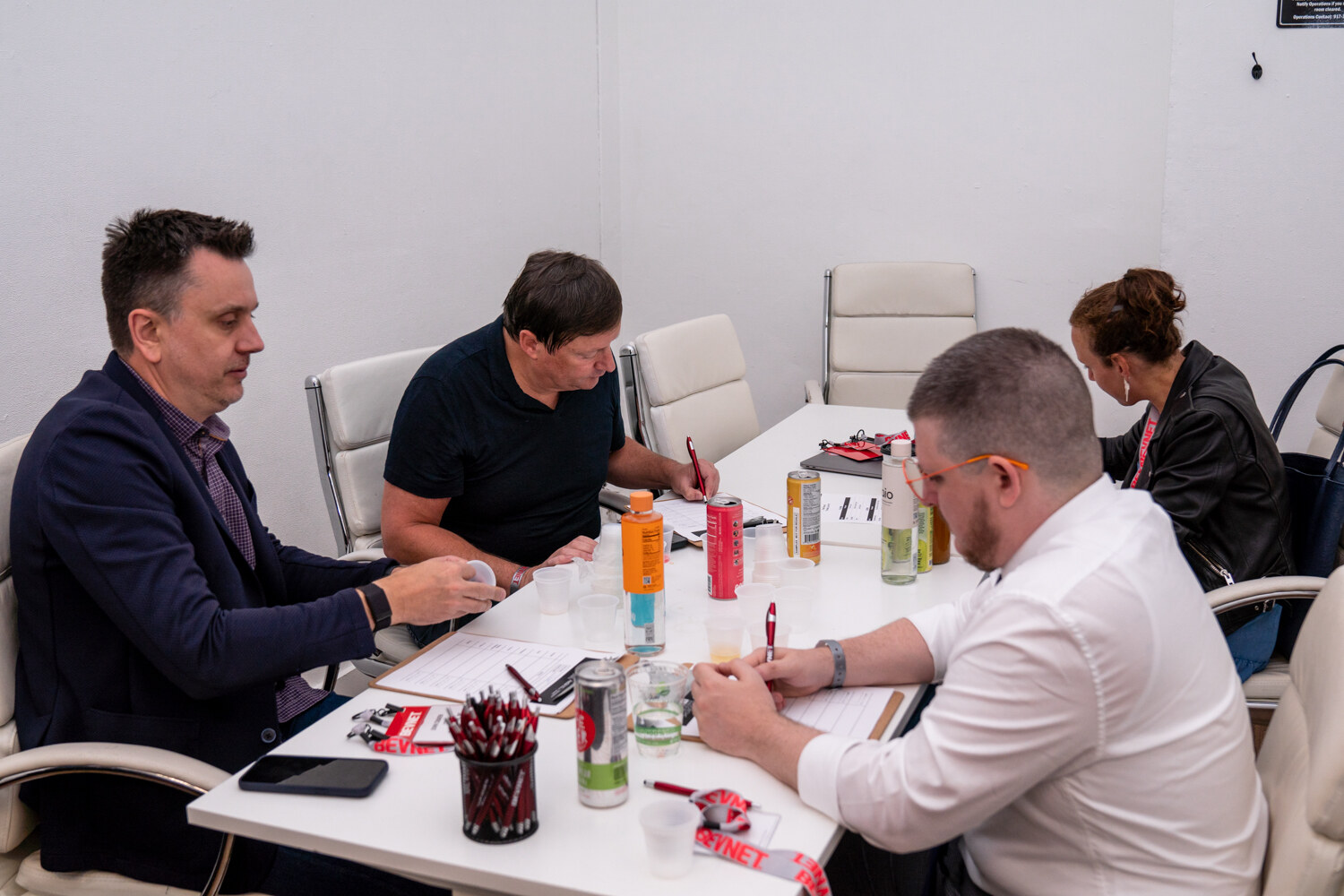 Four people sit around a white table covered with cans, bottles and pens, writing on score sheets in a small room.