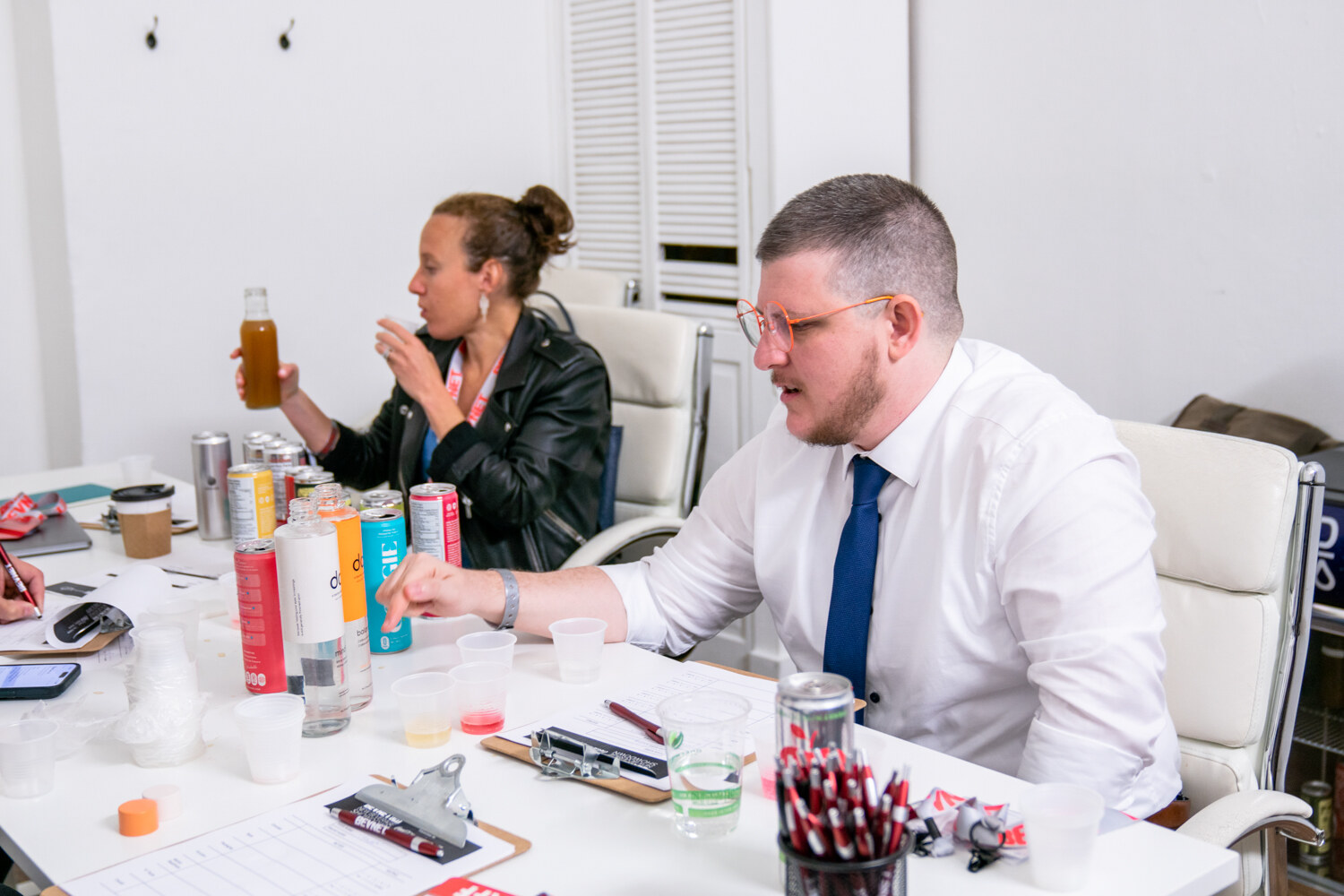 A woman in a black jacket and a man in a white shirt and blue tie lean over a table with cans, cups and clipboards.