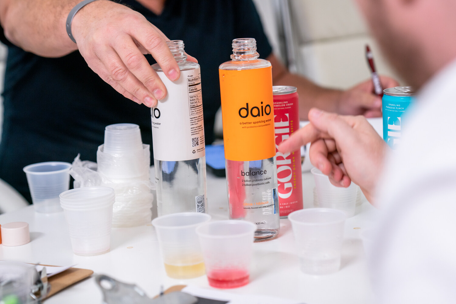 Close view of a table with bottles including an orange Daio bottle and a Gorgie can, small plastic tasting cups with samples, and hands reaching in.