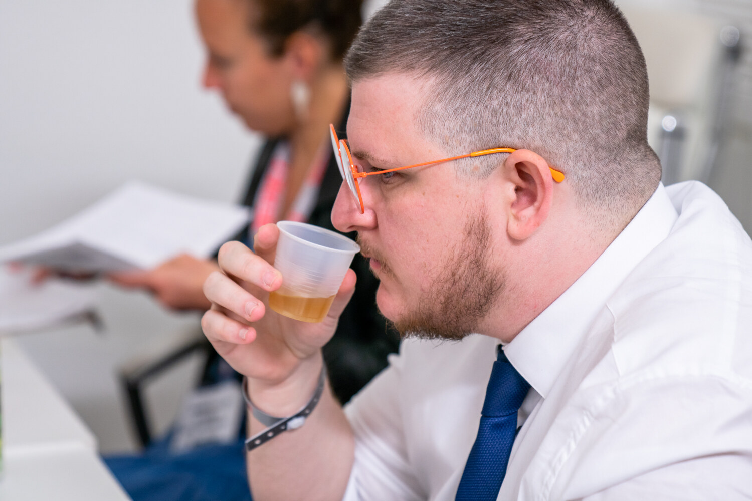 A man with orange-framed glasses and a trimmed beard in a white shirt and blue tie drinks from a small plastic cup while another judge reads papers behind him.