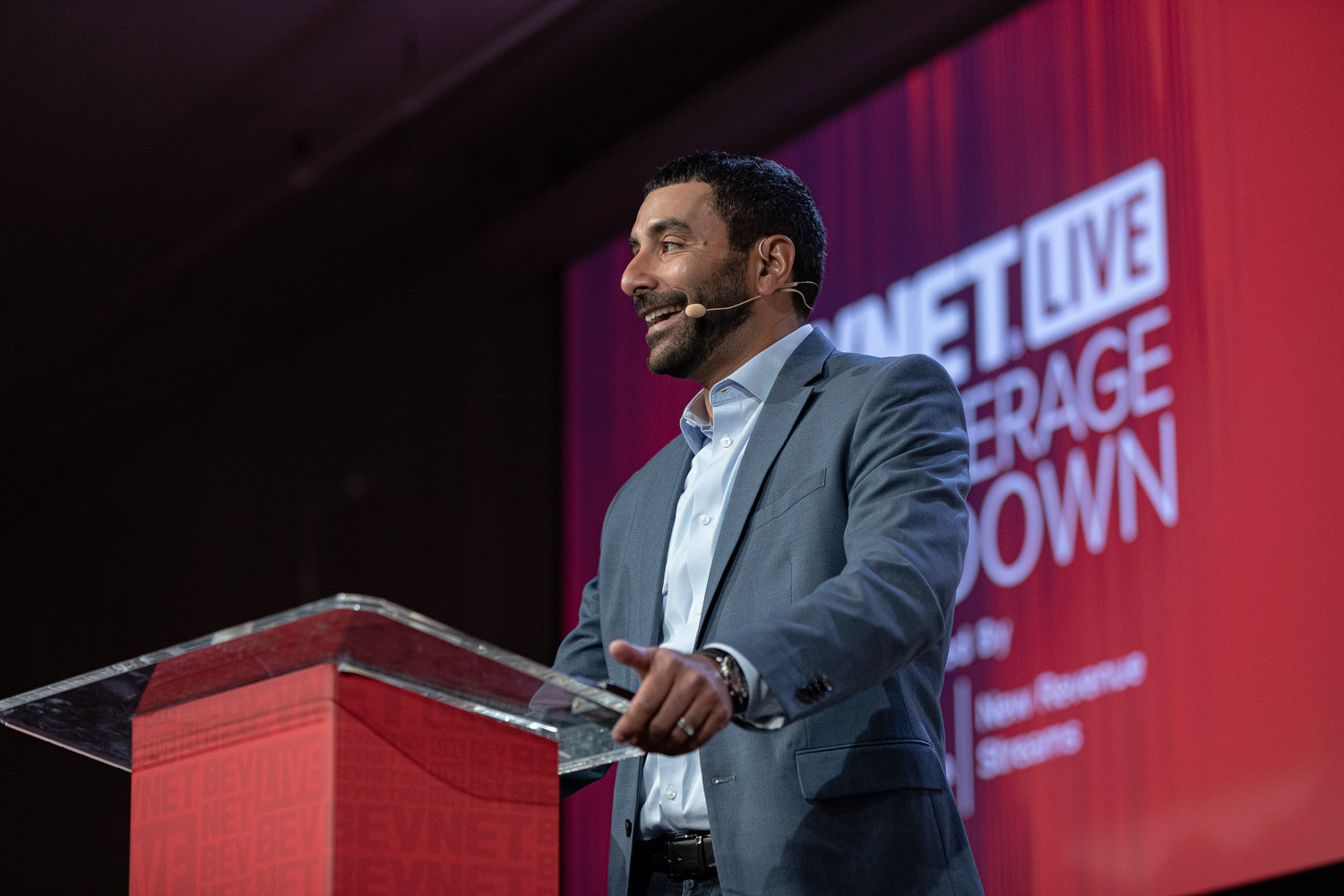 A man in a gray blazer and blue shirt speaks at a clear podium in front of a red screen that reads BevNET Live New Beverage Showdown.