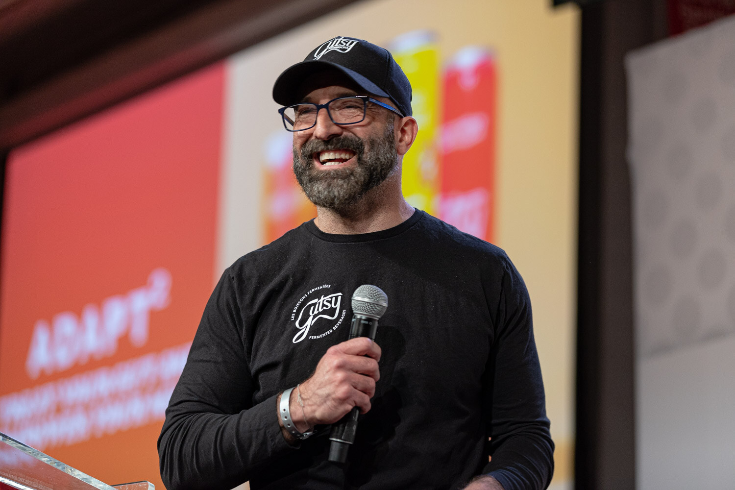 A smiling bearded man in a black cap and black shirt holds a microphone in front of an orange and red slide with the word Adapt.