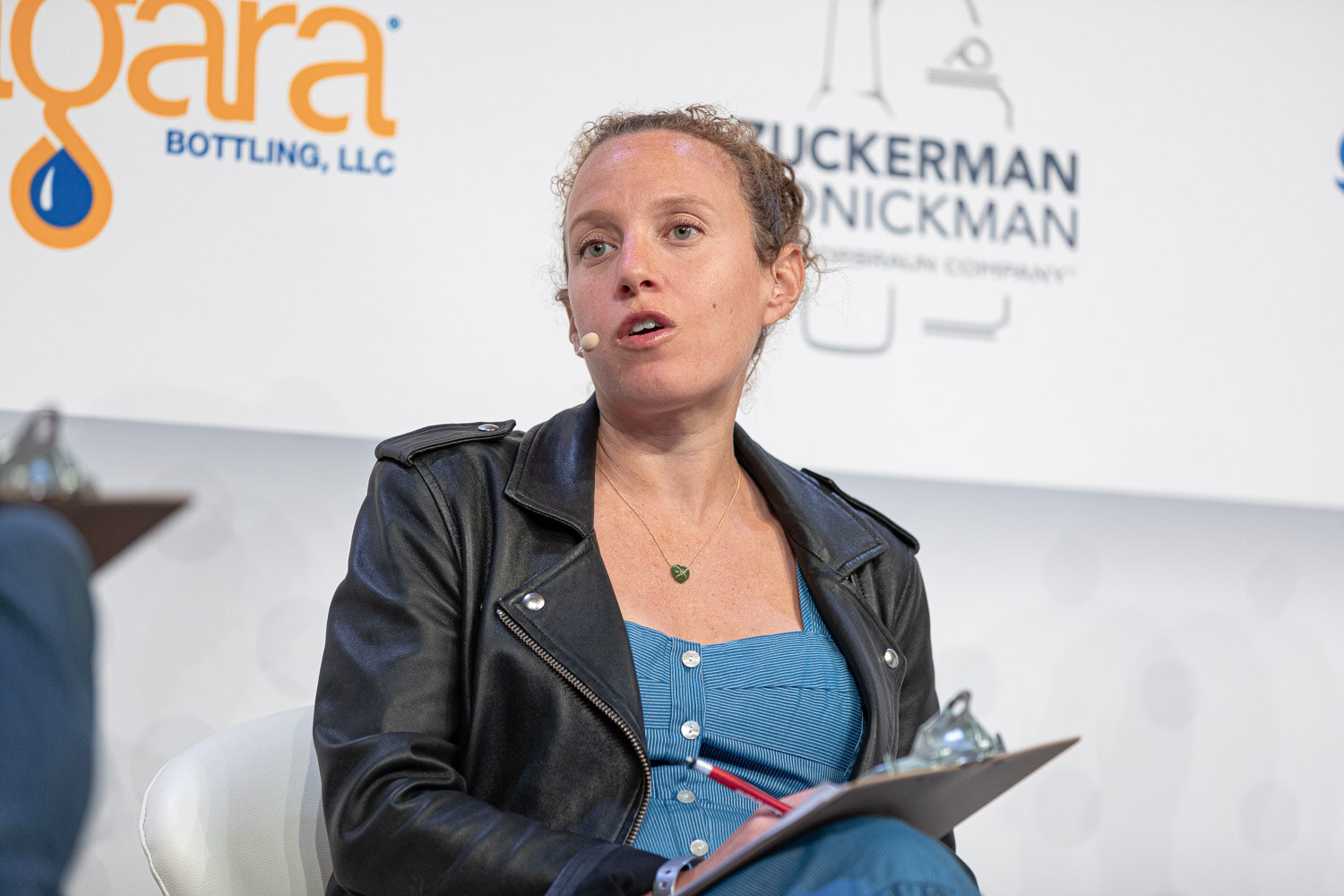 A woman in a black leather jacket and blue denim top sits with a clipboard in her lap in front of a Zuckerman Honickman sign.