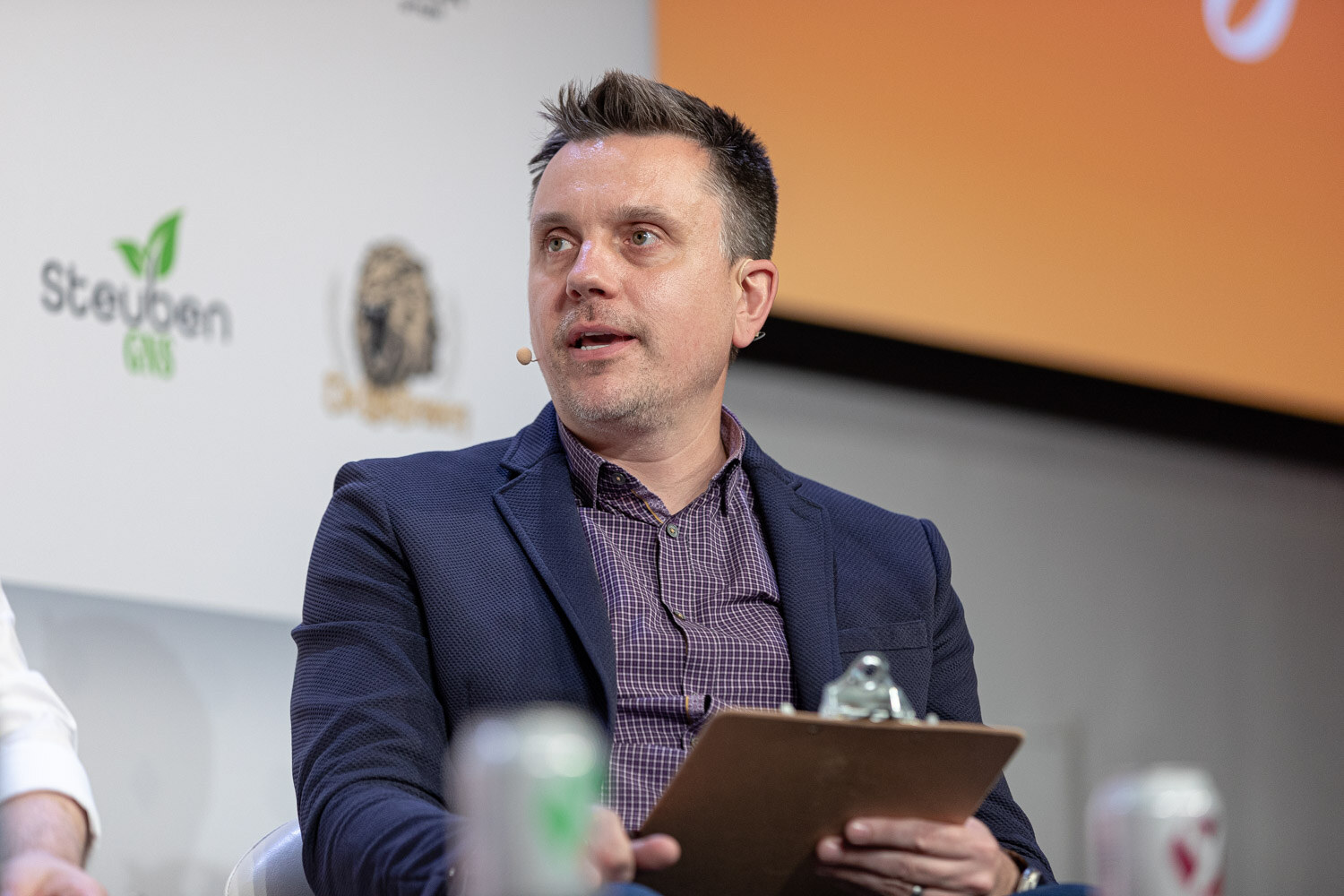 A man in a navy blazer and checked shirt holds a clipboard in front of a sponsor wall with an orange slide behind him.