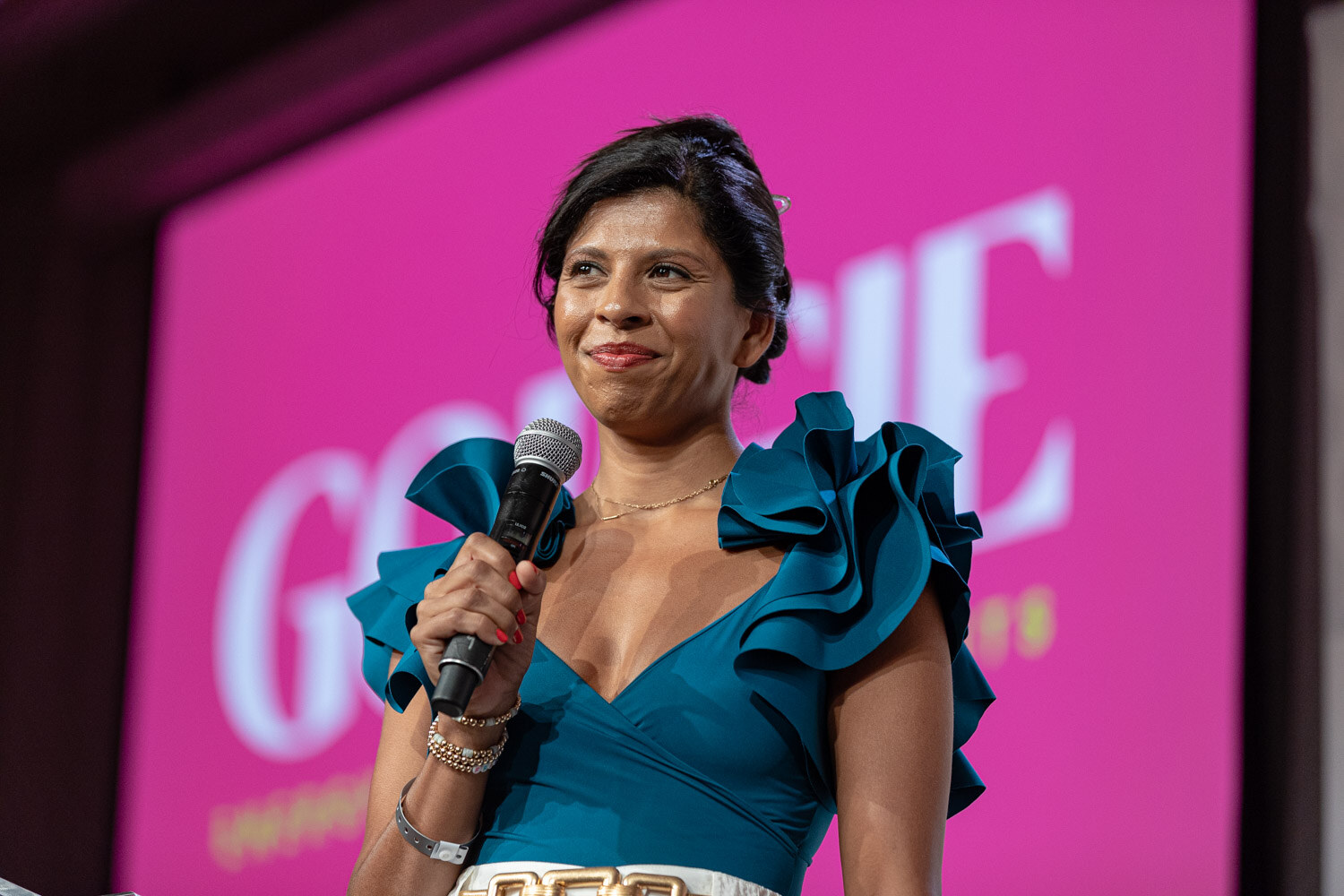 A woman in a teal ruffled top holds a microphone and smiles in front of a bright pink slide with the word Gorgie.