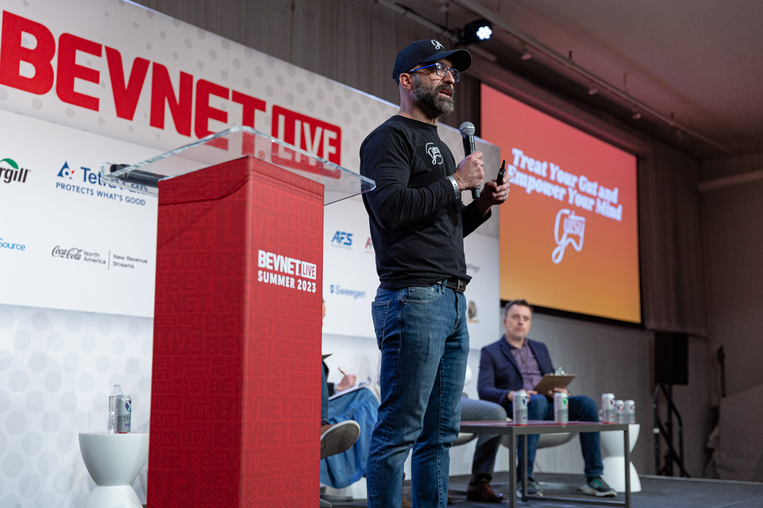 A man in a black shirt and navy cap stands beside a red BevNET Live podium with a screen at right reading Treat Your Gut and Empower Your Mind.