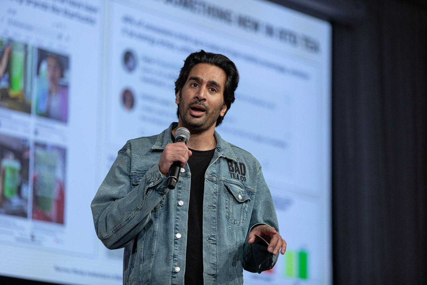 A man in a denim jacket over a black T-shirt holds a microphone in front of a projected slide of social media posts.