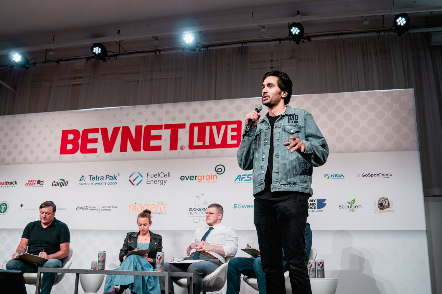 A man in a denim jacket stands on stage with a microphone while four judges sit on lounge chairs with clipboards beside him, under the BevNET Live logo.