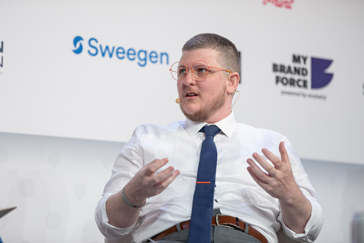 A man with glasses in a white shirt and navy tie holds both hands up while speaking, in front of a Sweegen and My Brand Force sponsor wall.