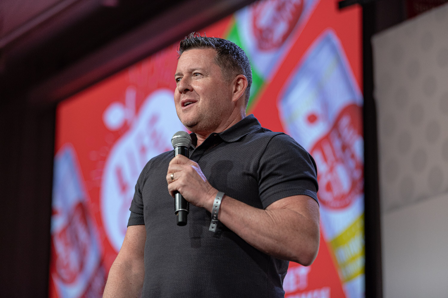 A man in a black polo shirt speaks into a microphone in front of a bright red slide with large illustrated cans.
