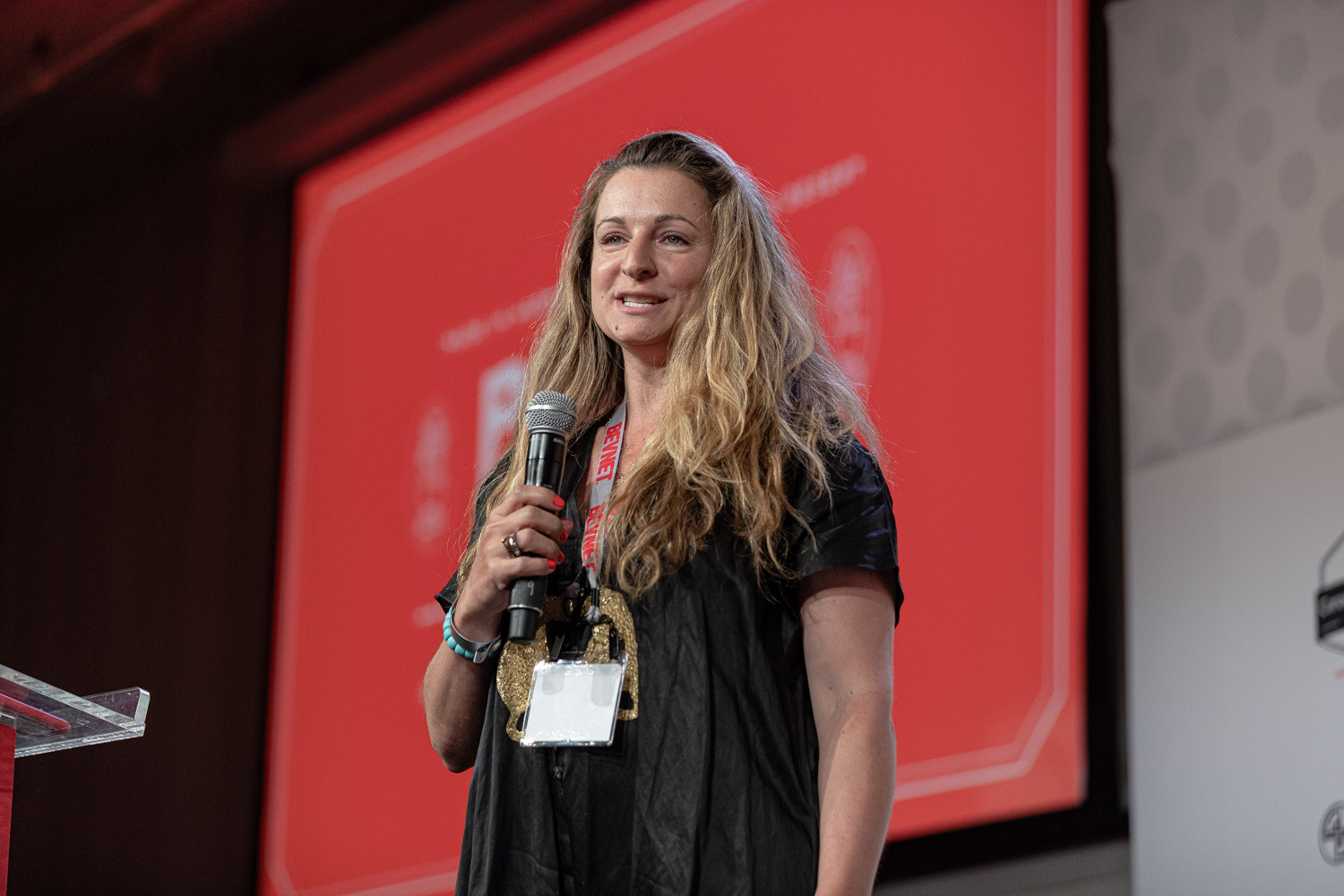A woman in a black dress with a lanyard and badge holds a microphone in front of a red projected slide.