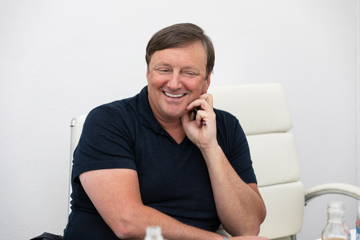 A smiling man in a navy polo shirt rests his hand near his ear while seated in a white chair.