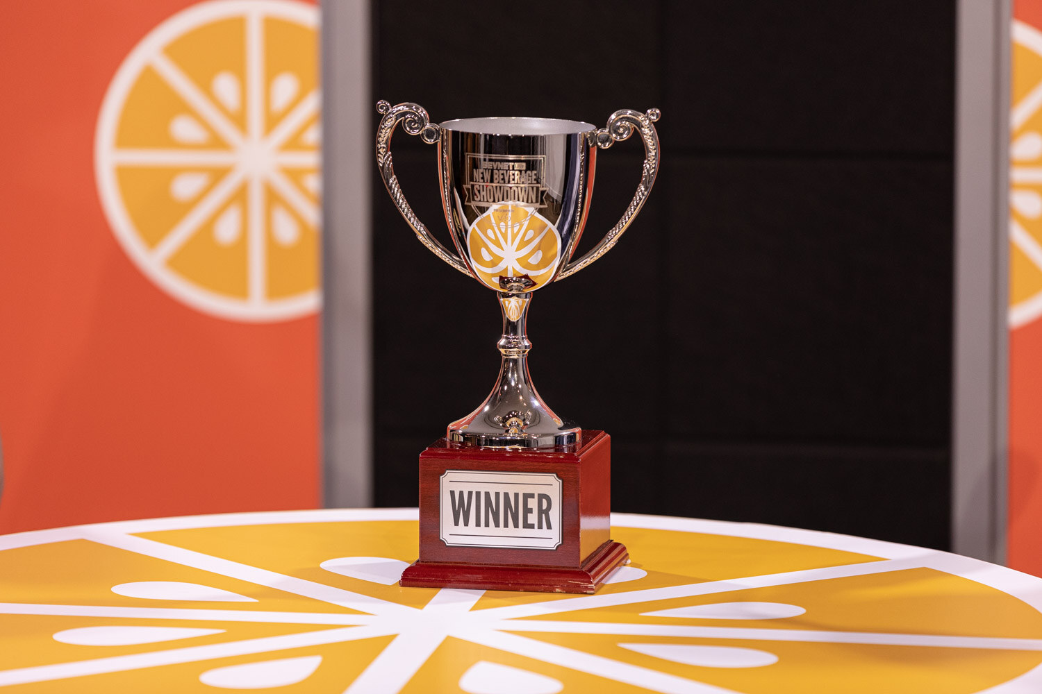A silver trophy with a plate reading Winner sits on a round orange-slice table with an orange Taste Radio backdrop behind it.
