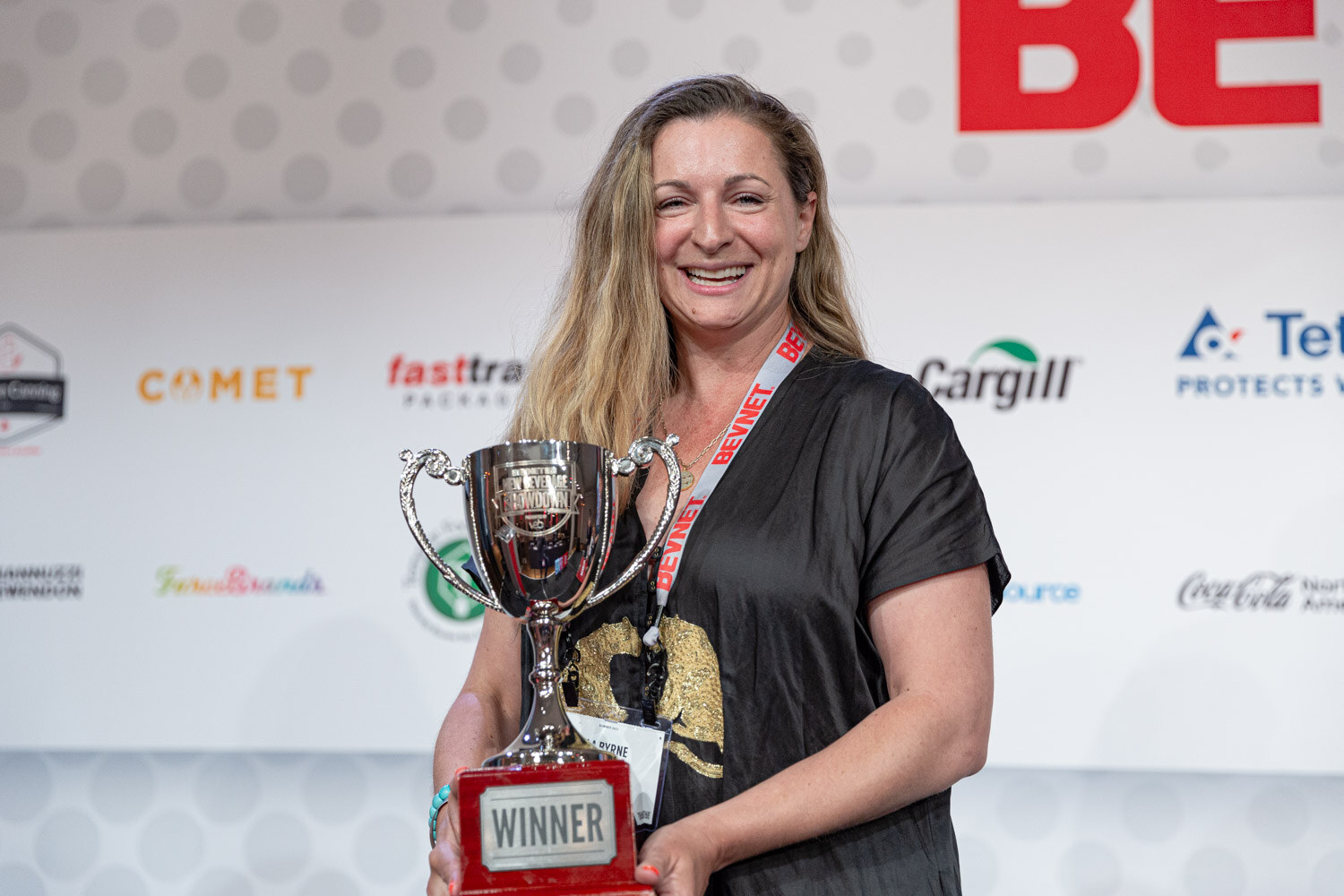 A smiling woman with long blonde hair and a lanyard holds a silver trophy marked Winner in front of a sponsor wall.