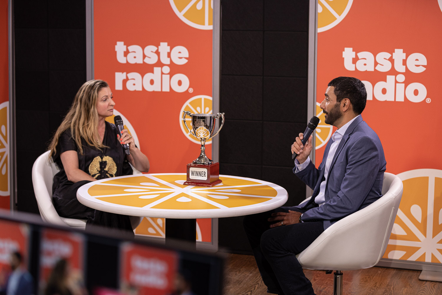 A woman in a dark top and a man in a gray blazer sit on either side of a round orange table with a trophy on it in front of an orange taste radio backdrop.