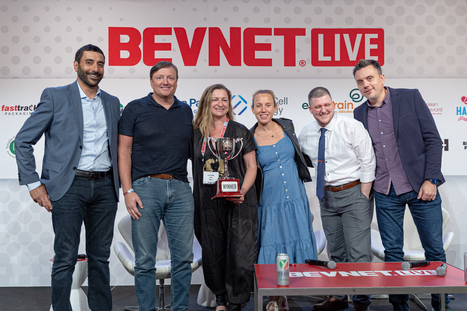 Six people in a row pose on stage with a silver trophy under the BevNET Live logo, with a red table in front of them.