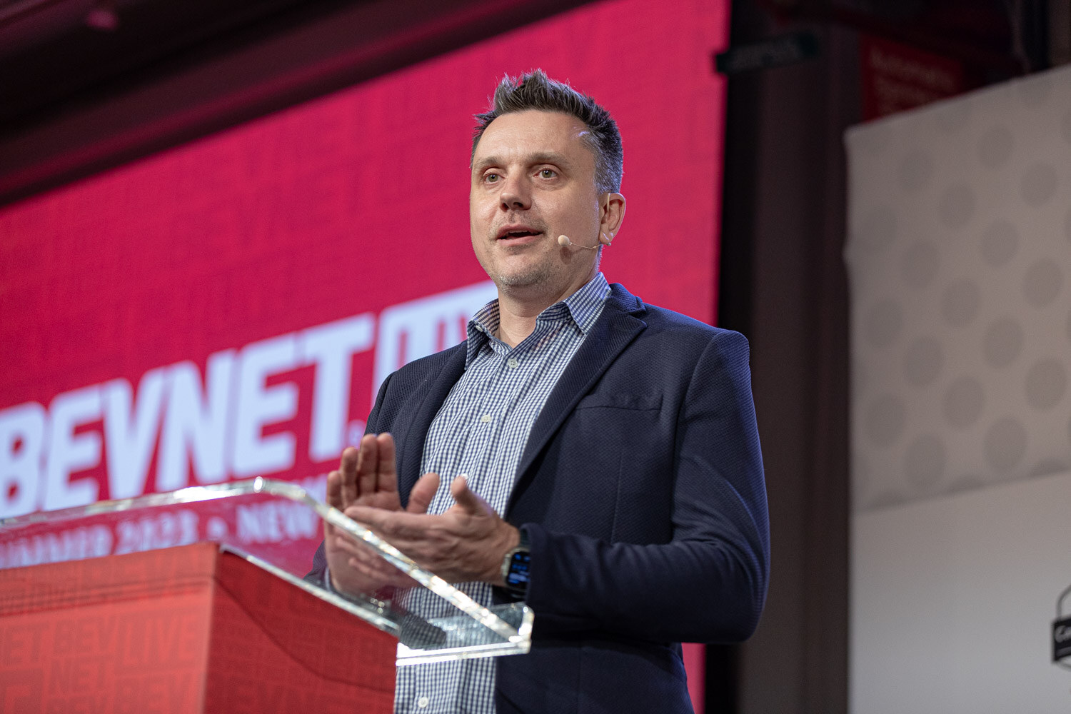 A man in a navy blazer and checked shirt gestures while speaking at a clear acrylic podium in front of a large red BevNET Live screen.