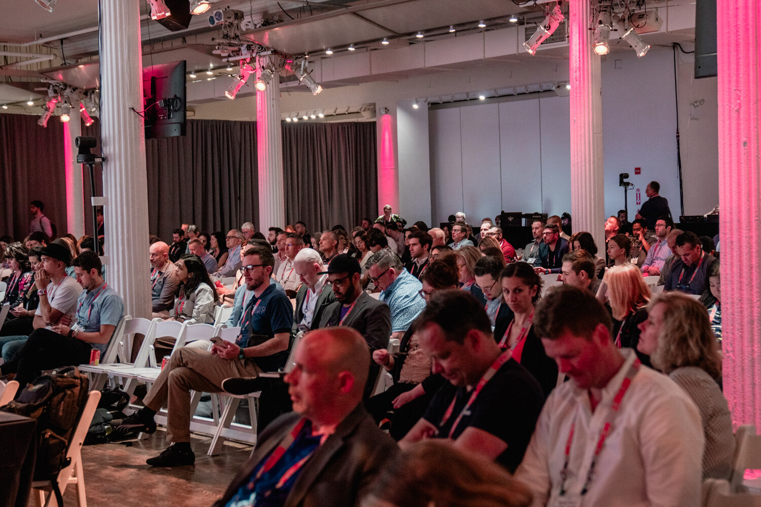 A crowded room of seated attendees wearing red lanyards watch a presentation in a loft space with pink-lit columns.