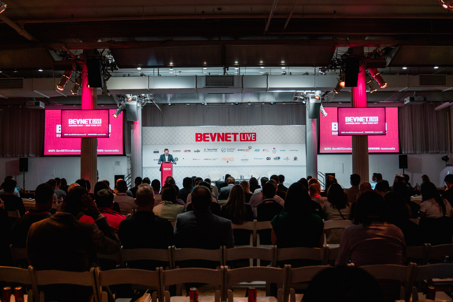 Wide view from the back of the main room showing seated attendees facing a stage with a red BevNET Live backdrop and two large screens.