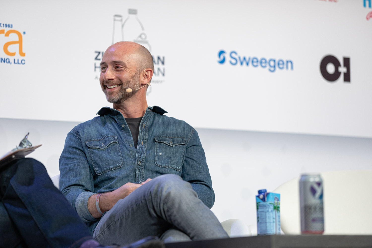A bald man in a denim shirt sits back with his legs crossed and smiles, with a Niagara-branded sponsor wall behind him.
