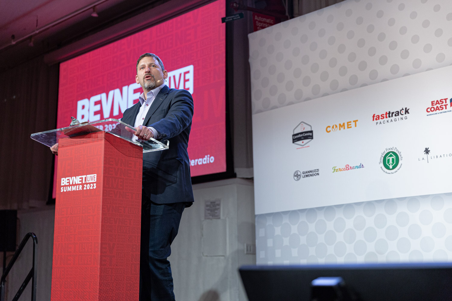 A man in a dark blazer stands behind a red podium marked BevNET Live Summer 2023 with a red screen and sponsor logos behind him.