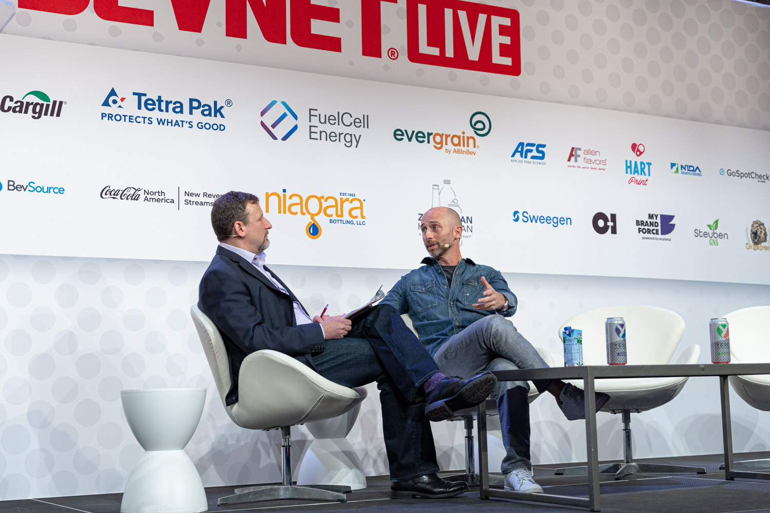 Wide shot of two men seated in white swivel chairs on stage in front of a white sponsor wall with a low table between them.