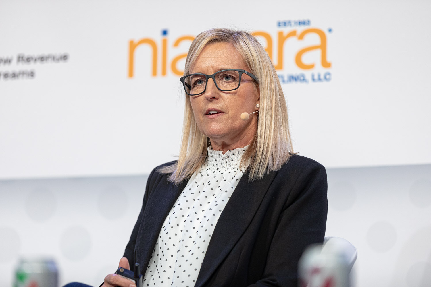 A woman with blonde hair and glasses in a polka-dot blouse and black blazer speaks in front of a Niagara sponsor sign.