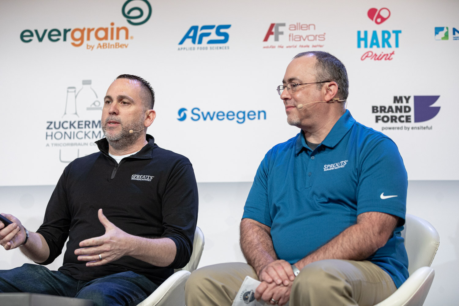 Two men seated on stage, one in a black quarter-zip and one in a blue polo shirt, both with small logos on the chest, in front of a sponsor wall.