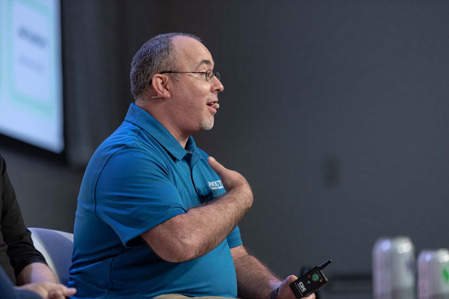 A man in a blue polo shirt and glasses speaks while holding a small remote in his hand against a dark gray background.